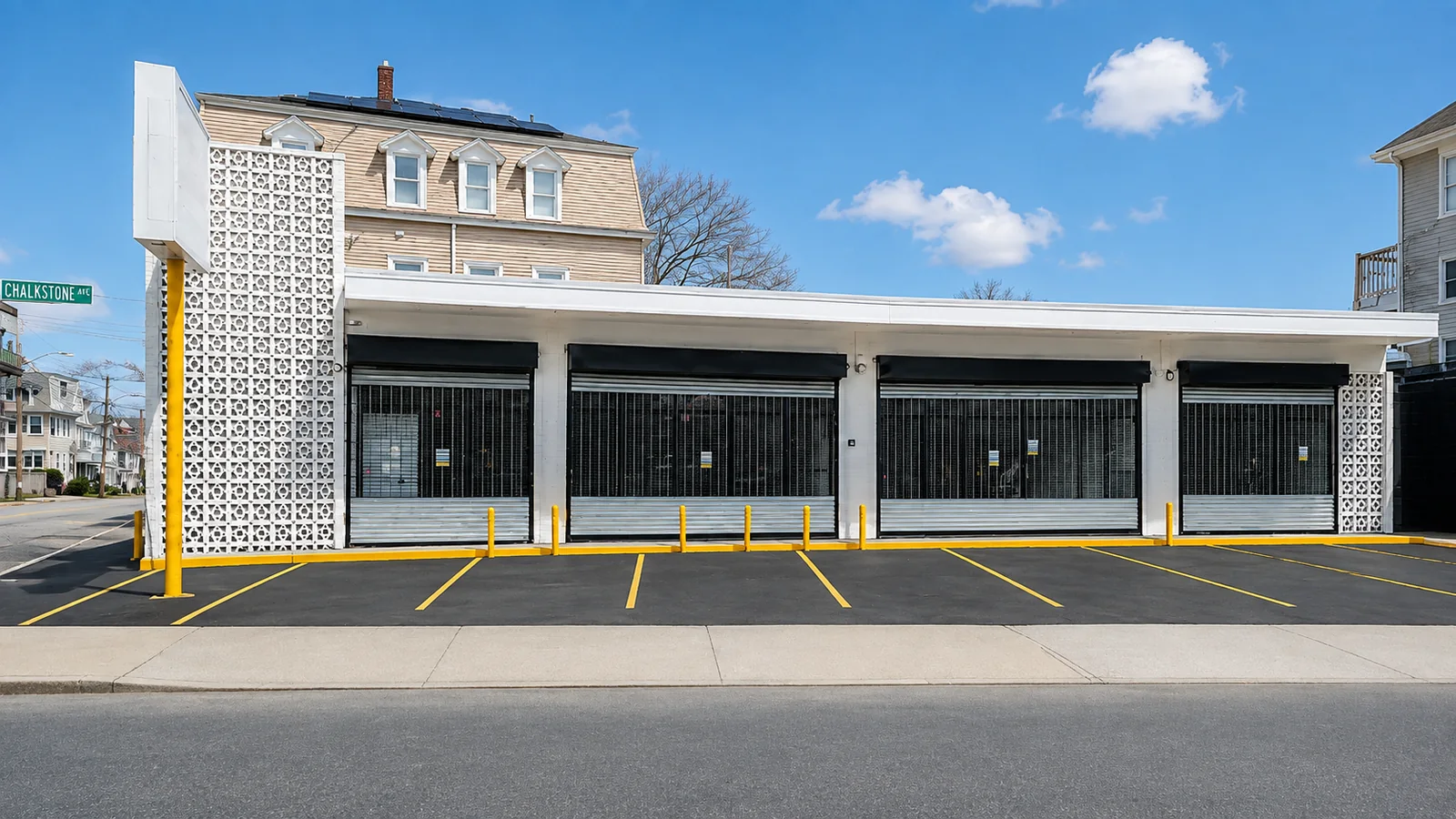 The front of 1115 Chalkstone Ave from Chalkstone Avenue: four storefronts under a canopy in a white one-story building, eight striped parking spaces out front, and the pylon sign at the corner of Chalkstone Ave and Home Ave